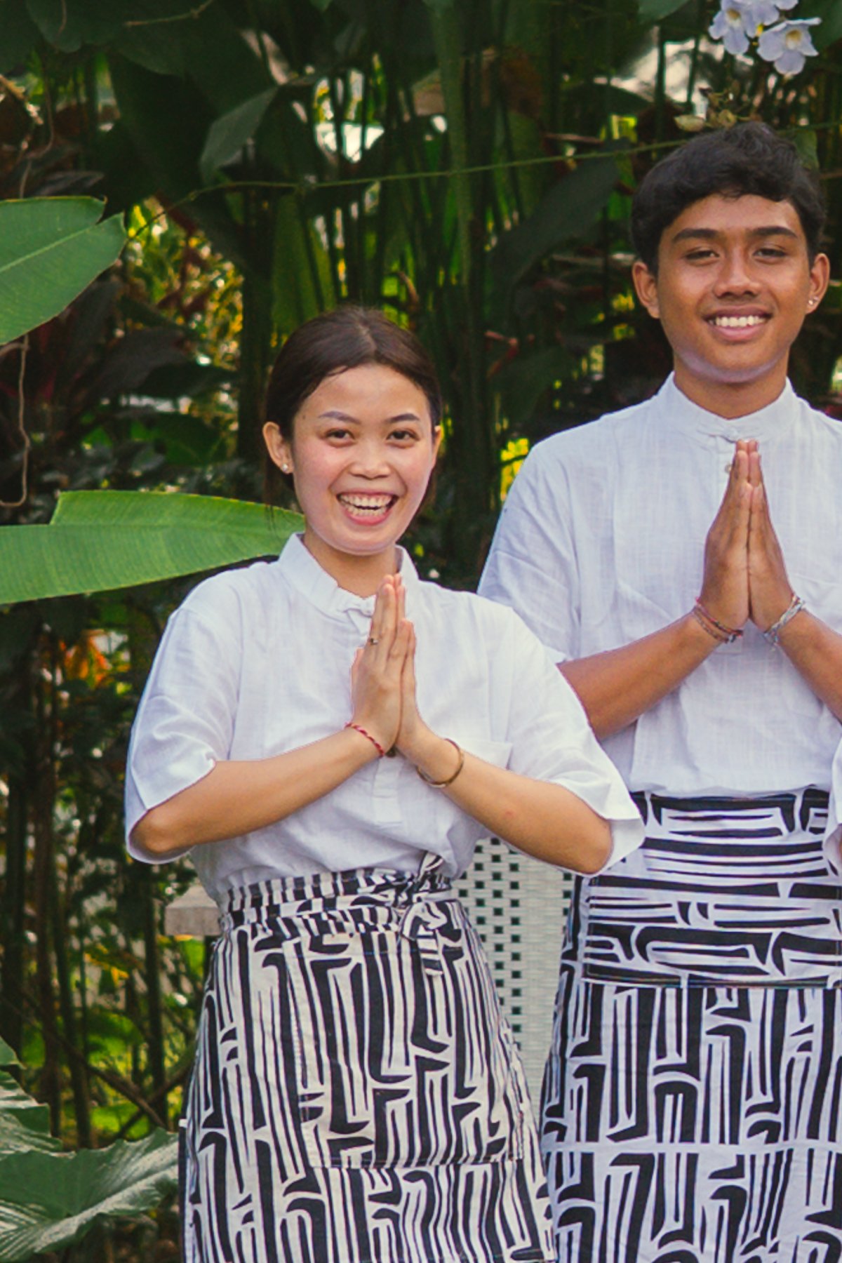 Two people smiling with hands pressed together in a prayer-like gesture, standing outdoors in front of lush green tropical plants.