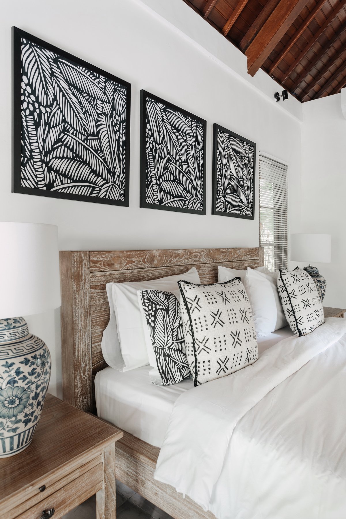 A bedroom with white walls, a wooden bed frame, white bedding, and decorative pillows with black and white patterns. Above the bed, there are three black-framed wall art pieces with black and white tropical leaf designs. On each side of the bed, there are bedside tables with white lamps and blue decorative vases. A window with white blinds is visible on the right side of the image.
