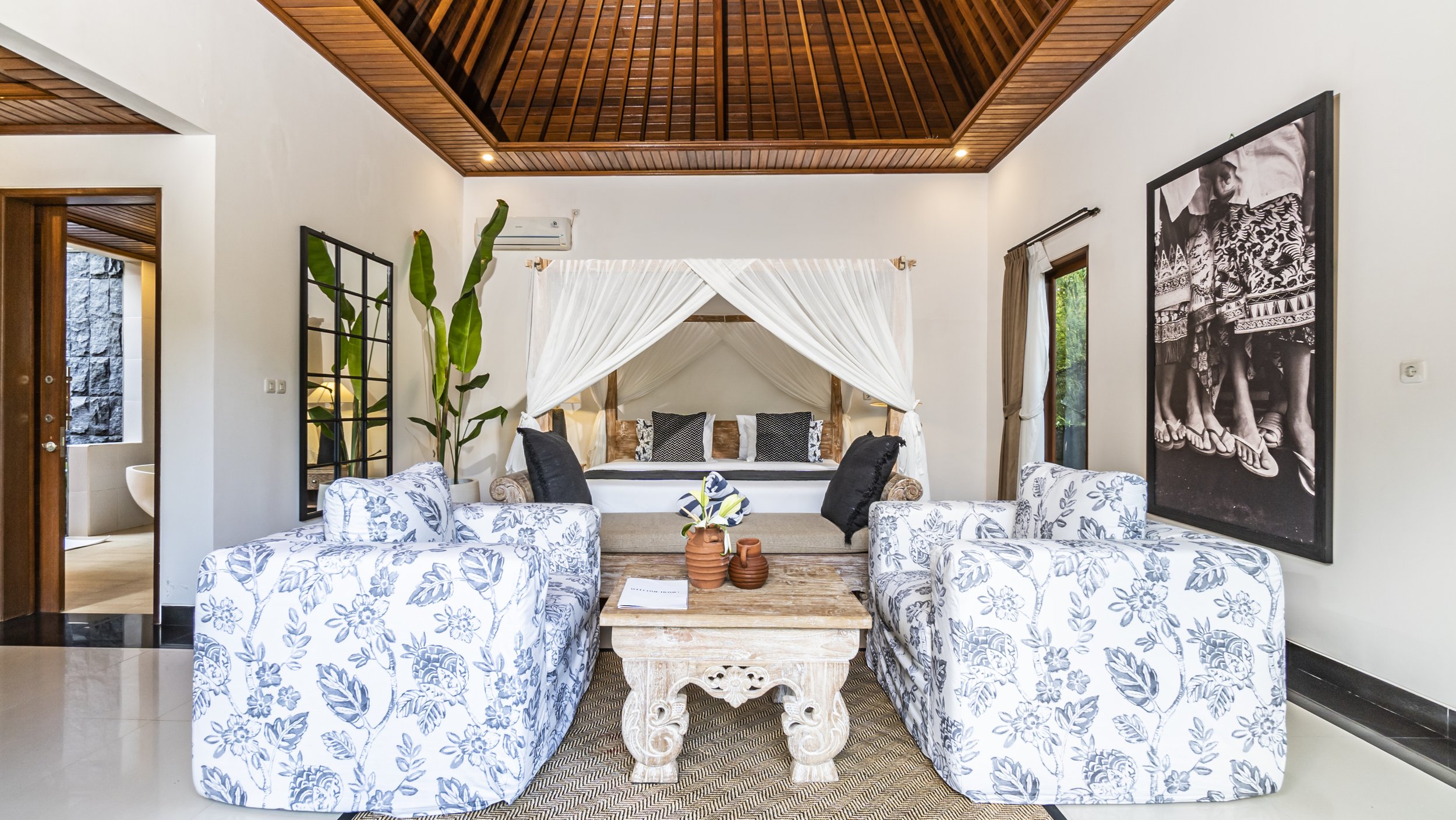 Living room with white walls, wooden ceiling, floral-patterned armchairs, a four-poster bed with drapes, and black-and-white artwork.
