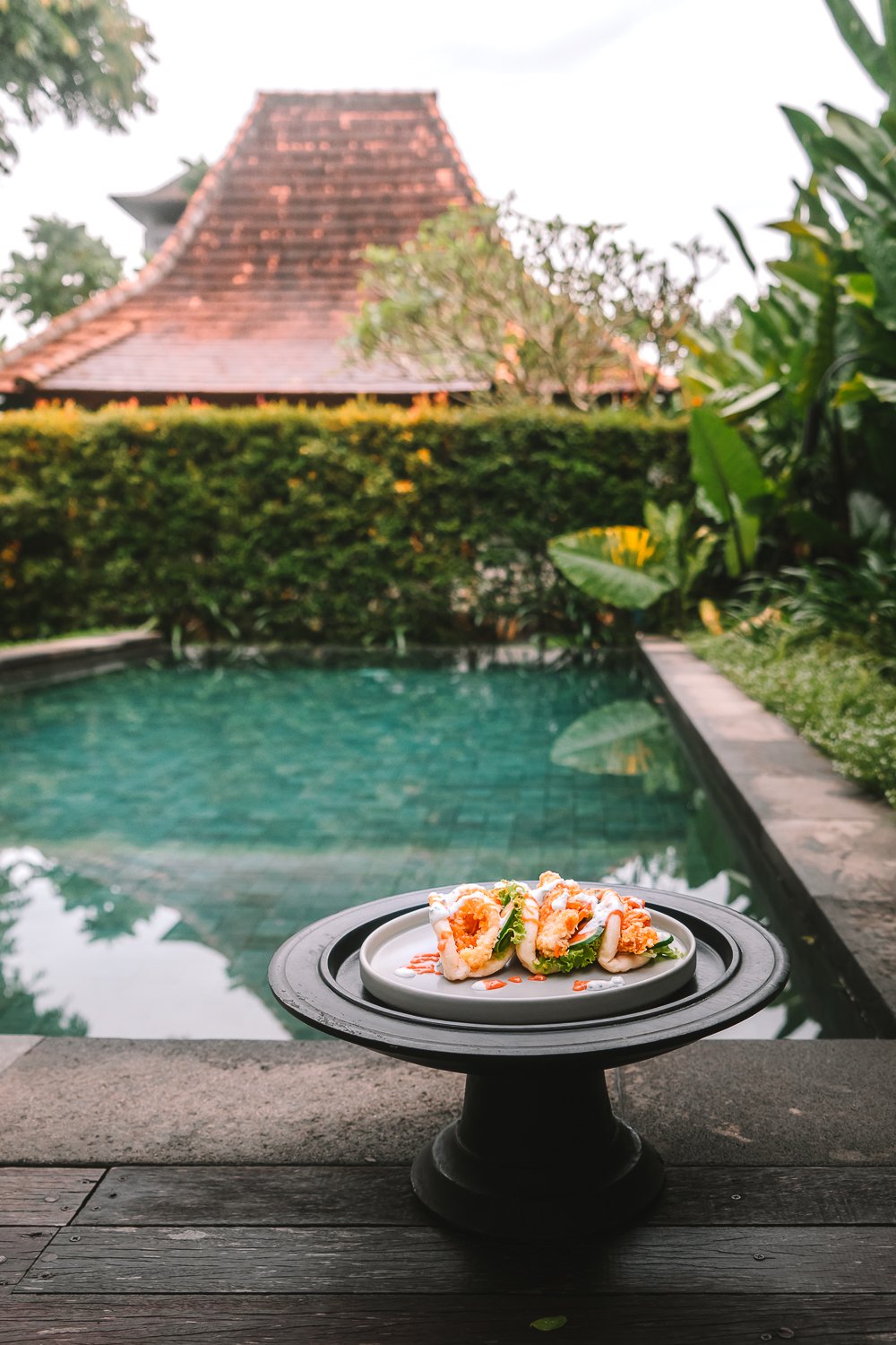 A plate of food on a stand, with a swimming pool, lush green bushes, and traditional thatched roof buildings in the background.