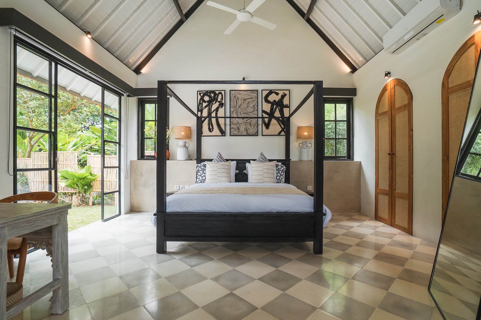 Modern bedroom with a four-poster black bed, large windows with greenery outside, and abstract art on the wall above the bed.
