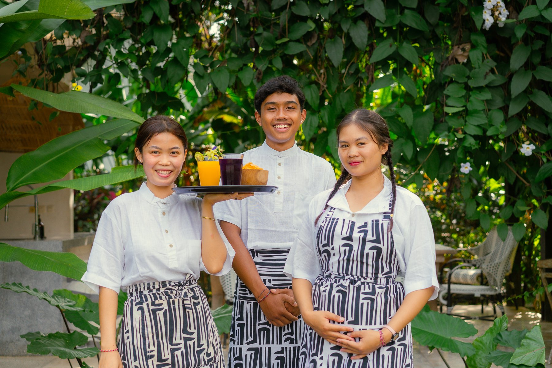 Three young people in white shirts and patterned aprons standing outdoors with lush green foliage, one holding a tray with drinks and desserts, smiling at the camera.