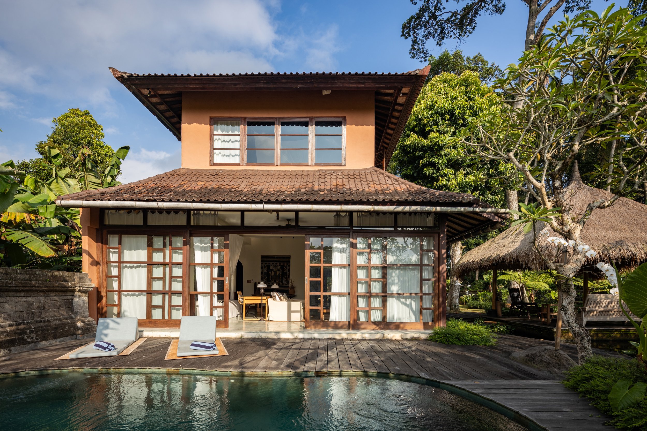 A tropical house with a tiled roof and large glass doors opens to a wooden patio with two lounge chairs by a small pool, surrounded by lush greenery and trees.