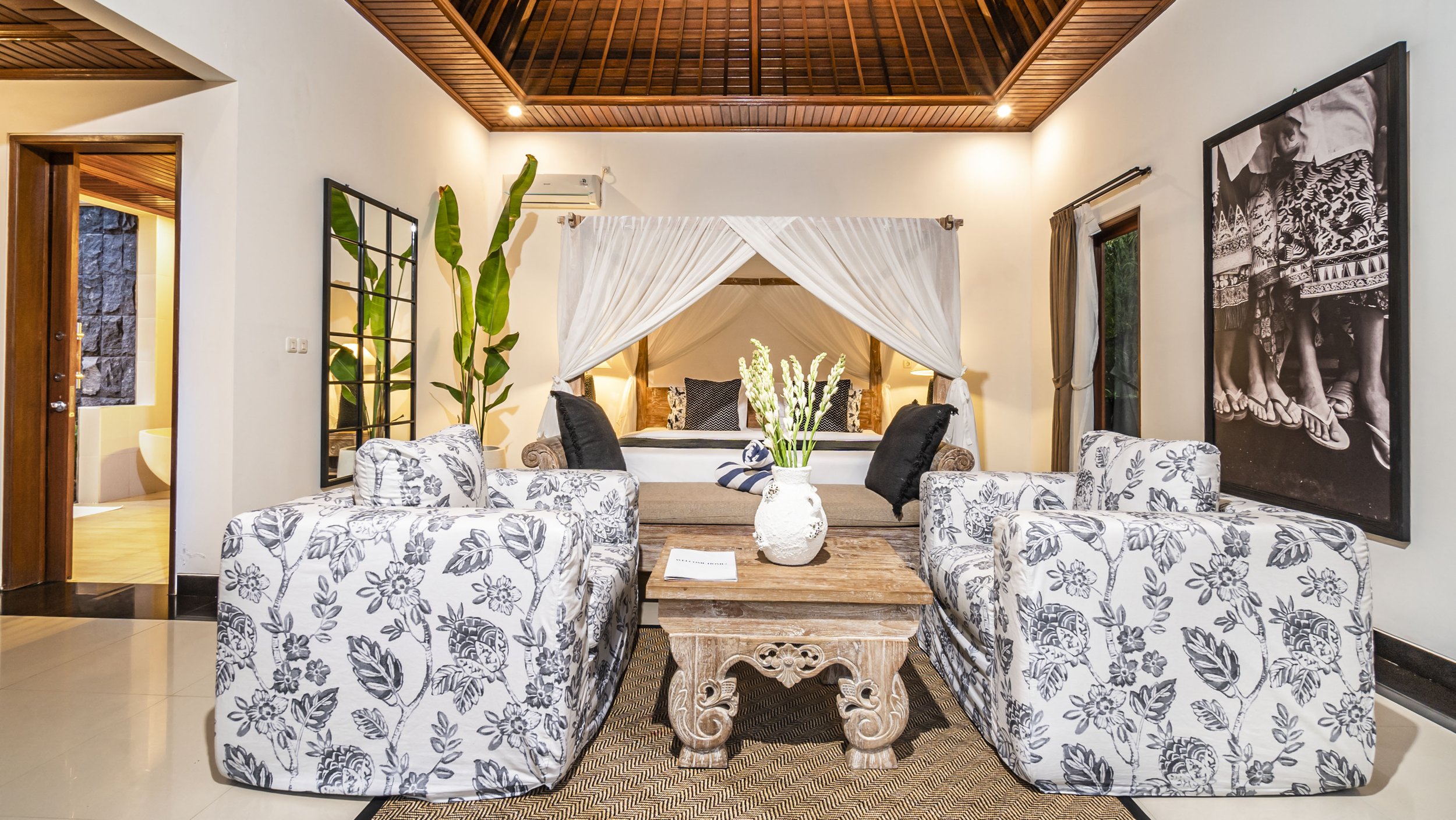 A tropical house with a tiled roof and large glass doors opens to a wooden patio with two lounge chairs by a small pool, surrounded by lush greenery and trees.