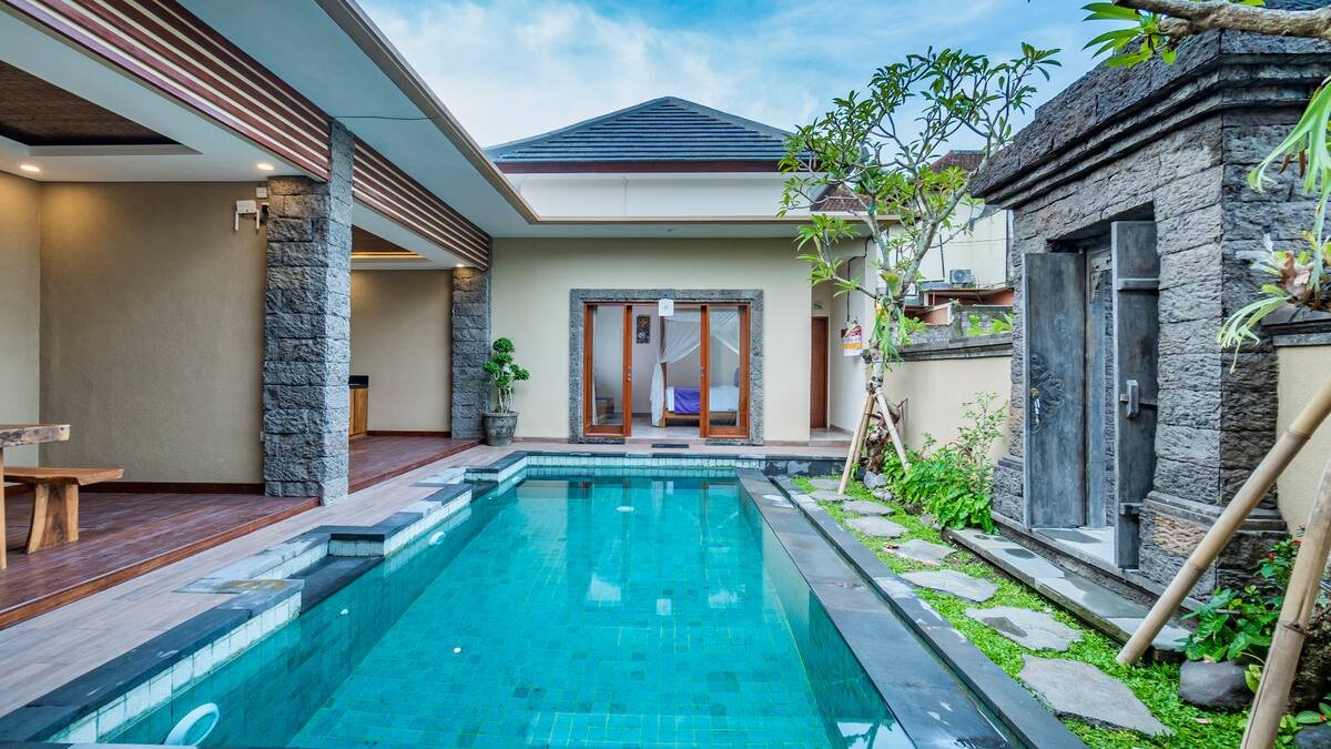 A tropical house with a tiled roof and large glass doors opens to a wooden patio with two lounge chairs by a small pool, surrounded by lush greenery and trees.