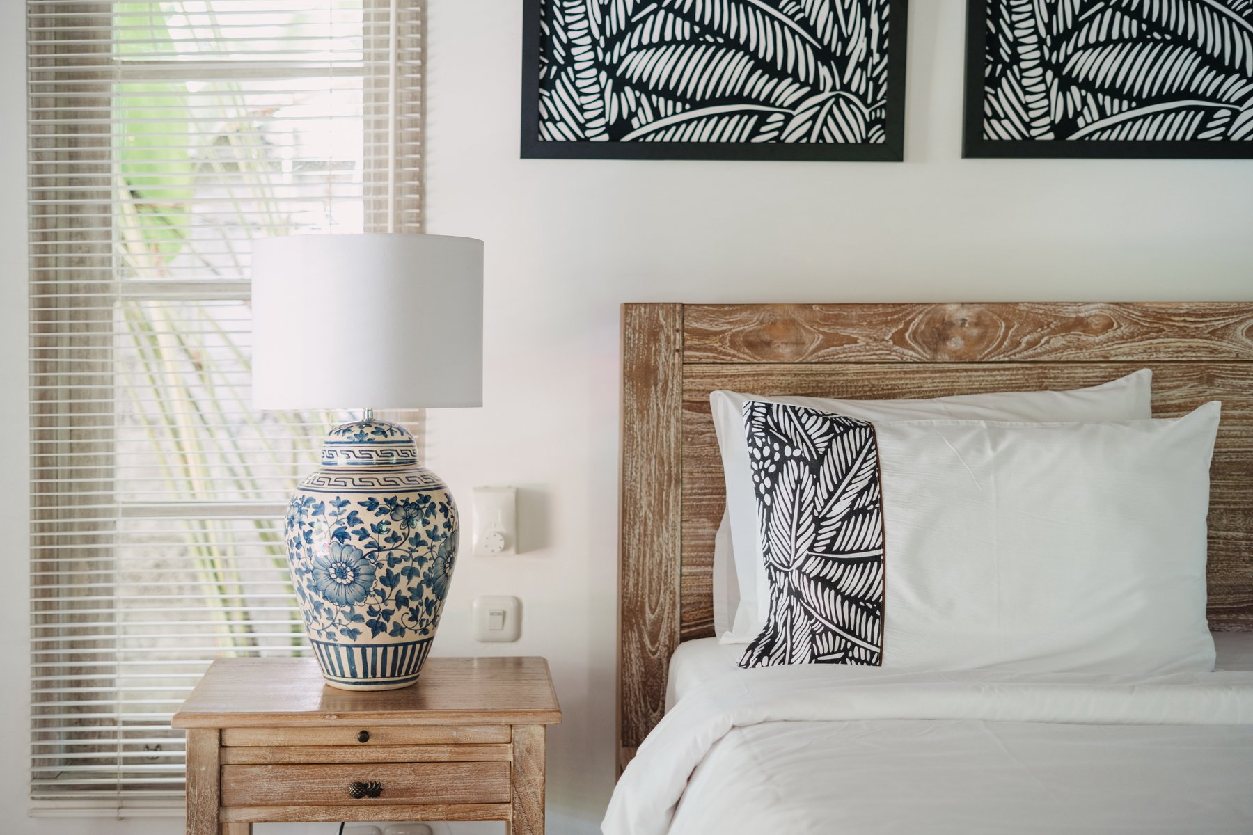 Bedroom with wooden headboard, white bedding with black and white decorative pillows, wooden nightstand with blue and white ceramic lamp, window with blinds, and framed black and white leaf art.