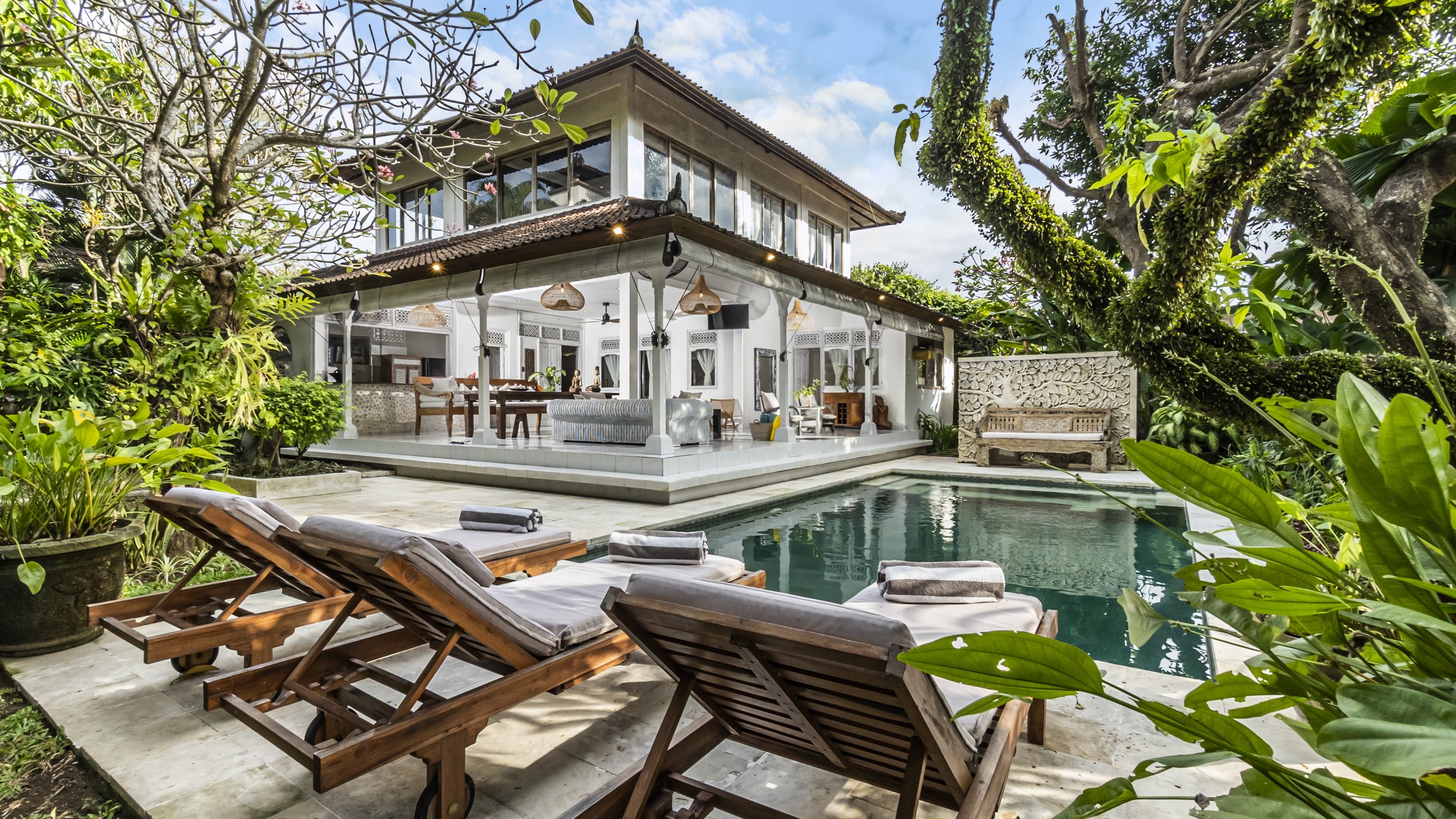 A tropical house with a tiled roof and large glass doors opens to a wooden patio with two lounge chairs by a small pool, surrounded by lush greenery and trees.
