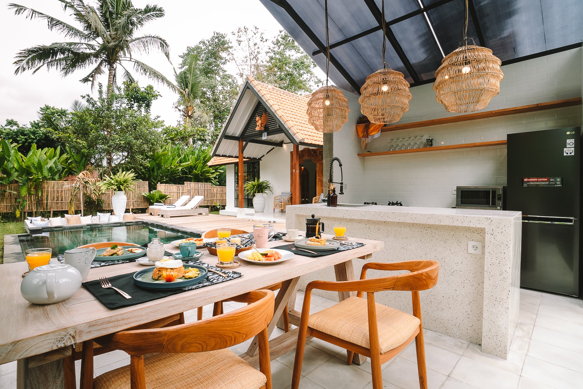 Outdoor dining area with breakfast setup, pool, and lush tropical garden. Wooden table with plates of food and glasses of orange juice, surrounded by wooden chairs. Poolside lounge chairs under a shaded area, tropical trees and plants in background.
