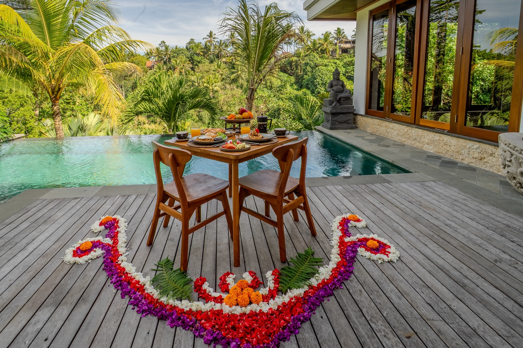 A tropical house with a tiled roof and large glass doors opens to a wooden patio with two lounge chairs by a small pool, surrounded by lush greenery and trees.