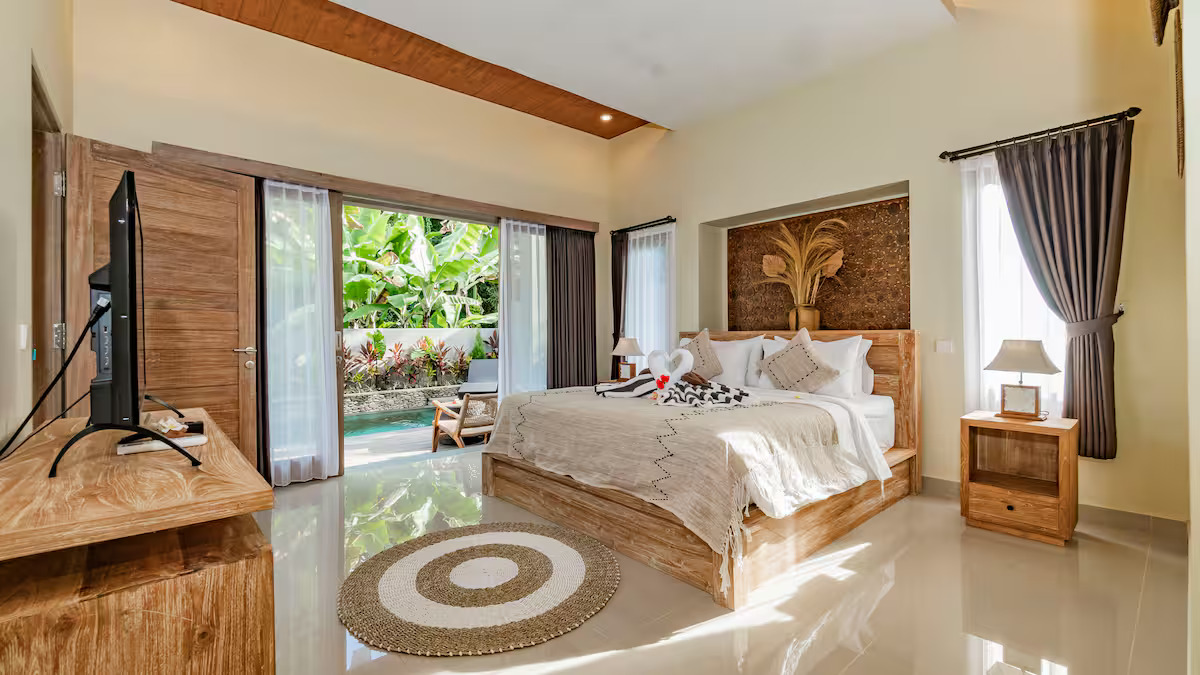A tropical house with a tiled roof and large glass doors opens to a wooden patio with two lounge chairs by a small pool, surrounded by lush greenery and trees.