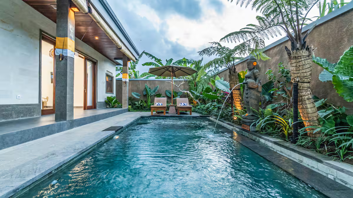 A tropical house with a tiled roof and large glass doors opens to a wooden patio with two lounge chairs by a small pool, surrounded by lush greenery and trees.
