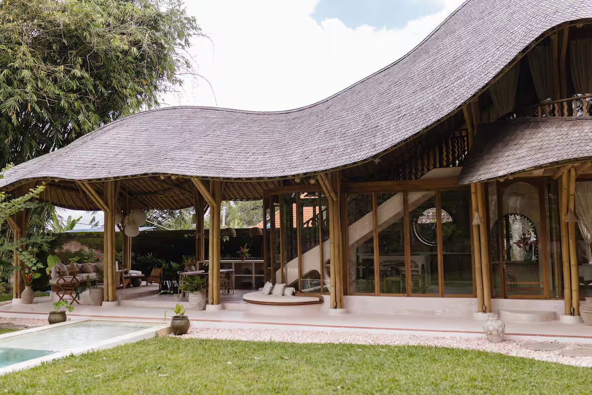 A tropical house with a tiled roof and large glass doors opens to a wooden patio with two lounge chairs by a small pool, surrounded by lush greenery and trees.